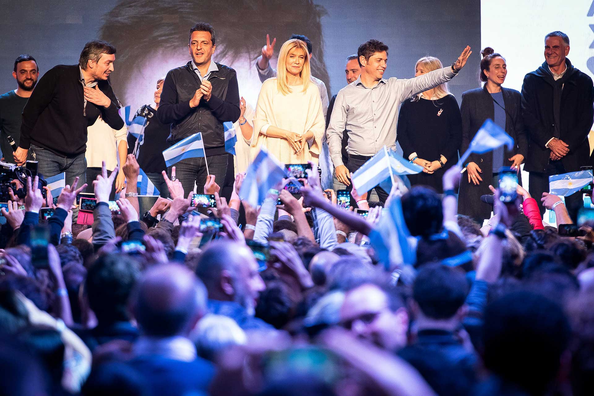 Fotografía de los candidatos electos por el 'Frente de Todos' festejando el triunfo en la campaña electoral 2019. En primer plano la gente festejando dentro del búnker y de fondo los candidatos electos festejando en el escenario