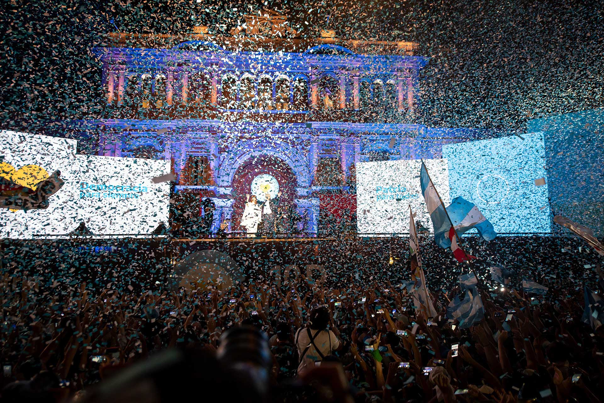 Fotografía de Alberto Fernández y Cristina Fernández de Kirchner ingresando al escenario para los festejos en la puerta de 'Casa Rosada. Componen la fotografía una multitud en la plaza, una neblina de papelitos al aire y de fondo el Presidente y Vicepresidenta electos sobre el escenario montado frente a la 'Casa Rosada'