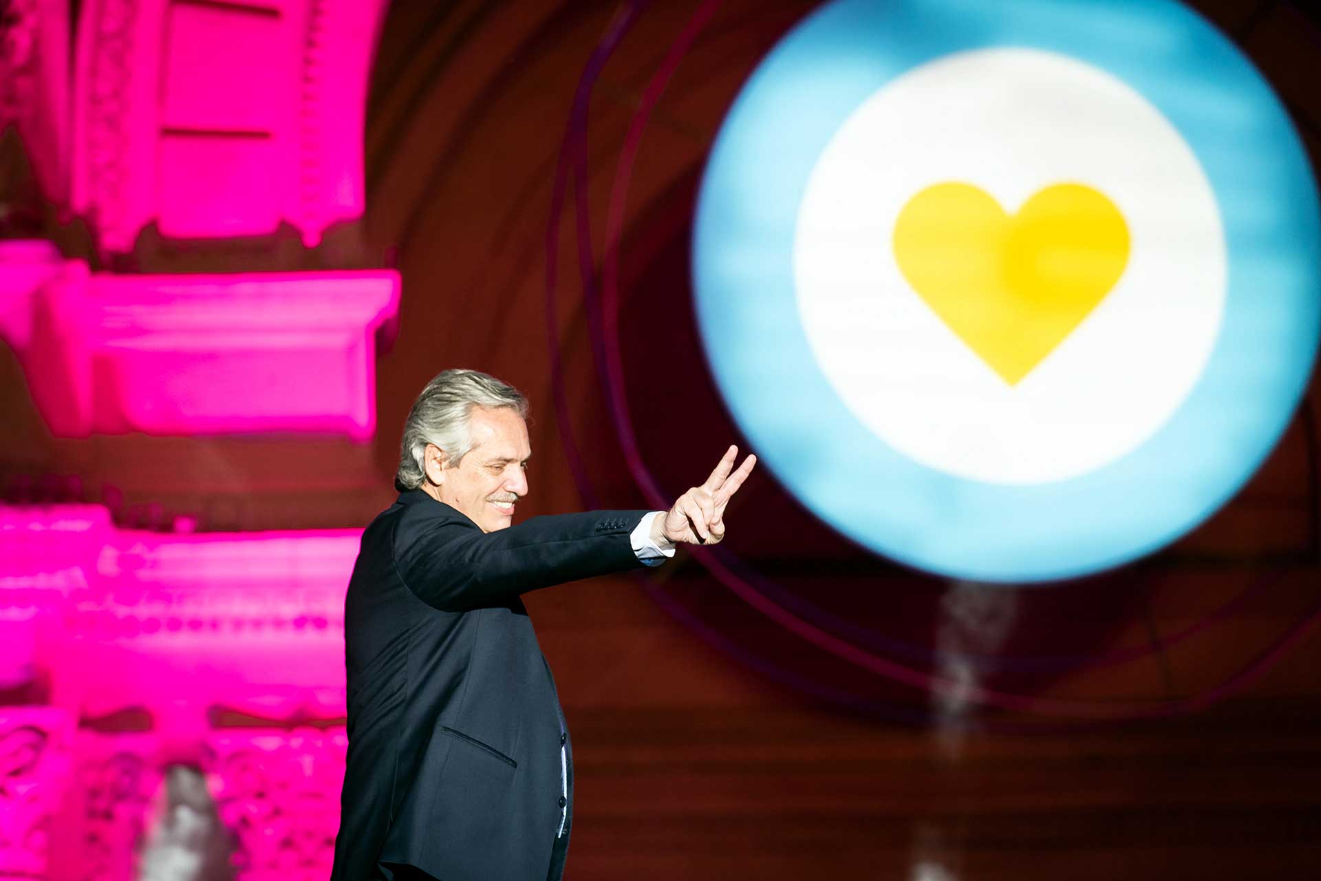 Fotografía de Alberto Fernández saludando al público con su brazo extendido y los dedos de su mano formando la 'V' de la Victoria. De fondo una imagen, proyectada en una pantalla gigante, de una escarapela argentina con un corazón en el centro