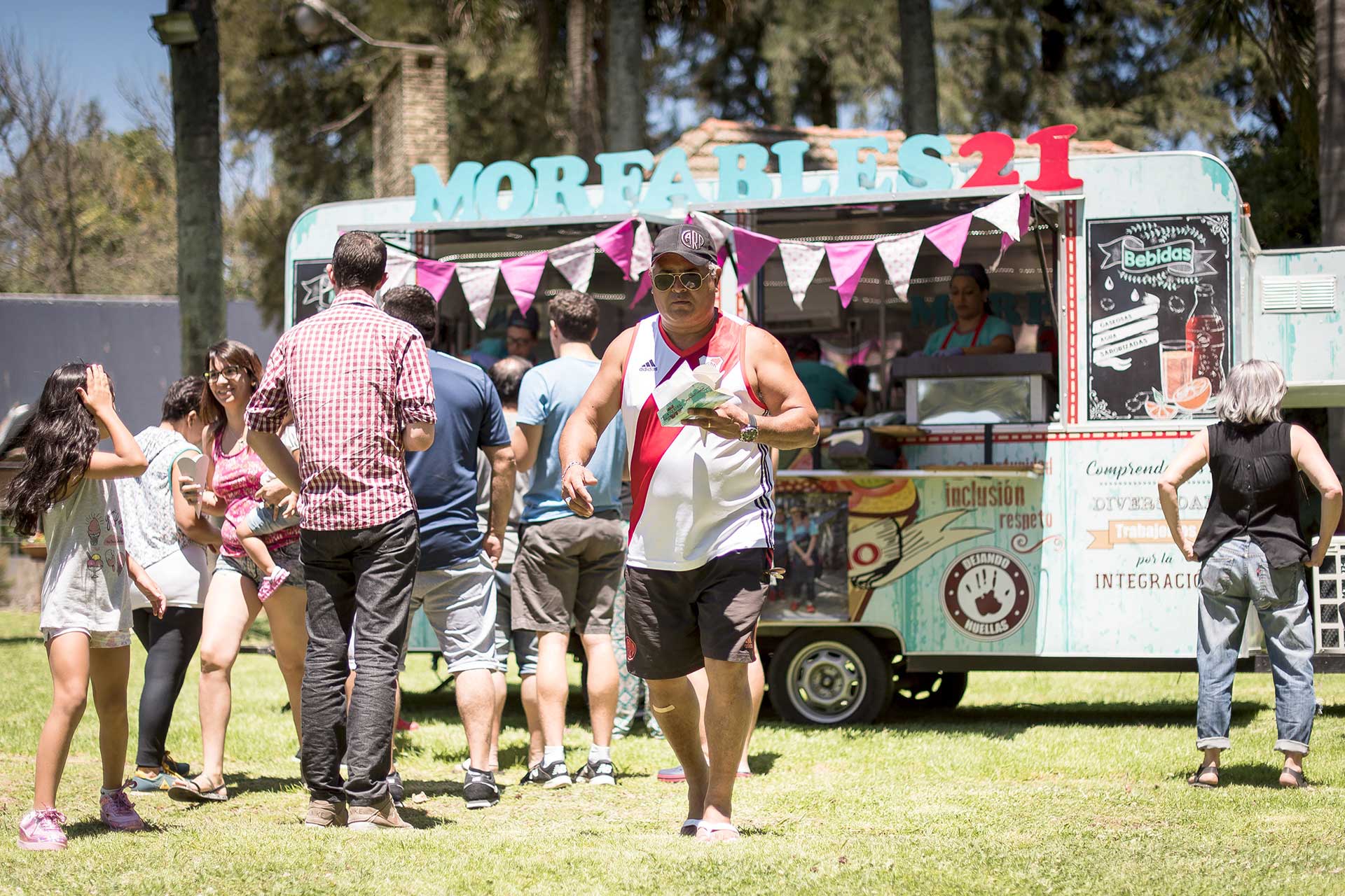 Fotografía de un hombre caminando con un cono de papas fritas. De fondo un foodtruck gastronómico, con una importante fila de gente, parte del evento, matutino, organizado por la empresa 'Danone Argentina'