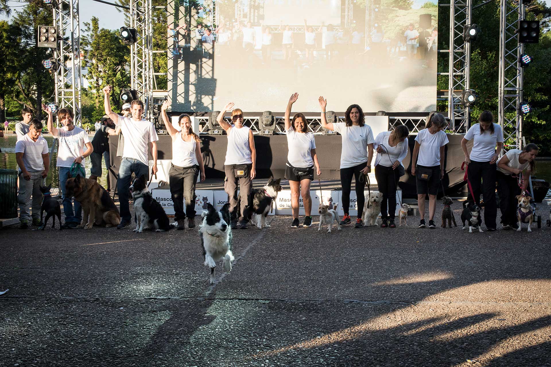 Fotografía de once participantes, frente al escenario, junto a sus mascotas, delante de ellos un perro corriendo en primer plano. Cobertura del evento 'Mascotas de la Ciudad', organizado por el Gobierno de la Ciudad Autónoma de Buenos Aires