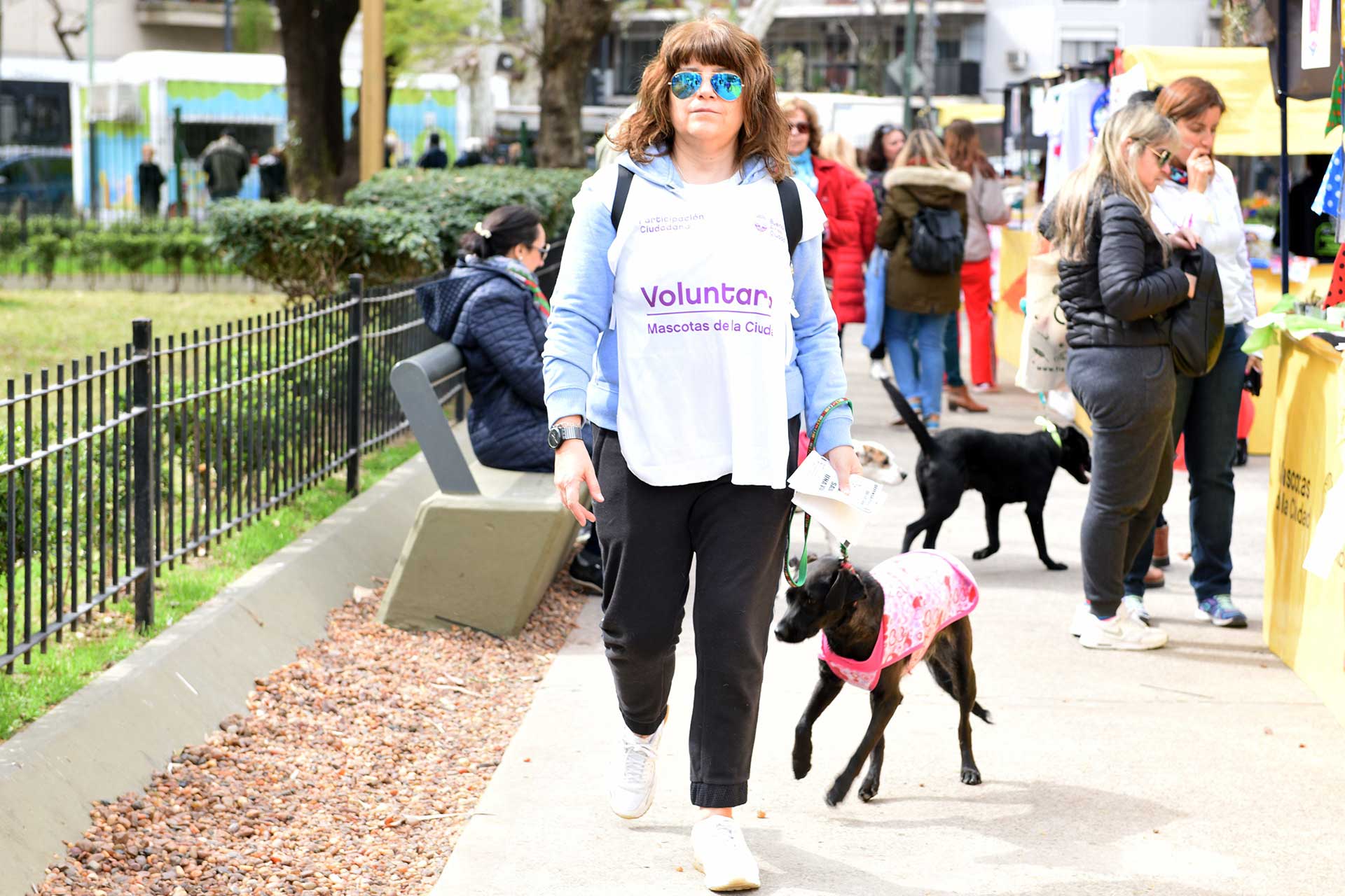 Fotografía de una mujer paseando a su perro mientras recorre los stands dentro del evento, de fondo gran parte del público, disfrutando de los mismos. Cobertura del evento 'Mascotas de la Ciudad', organizado por el Gobierno de la Ciudad Autónoma de Buenos Aires