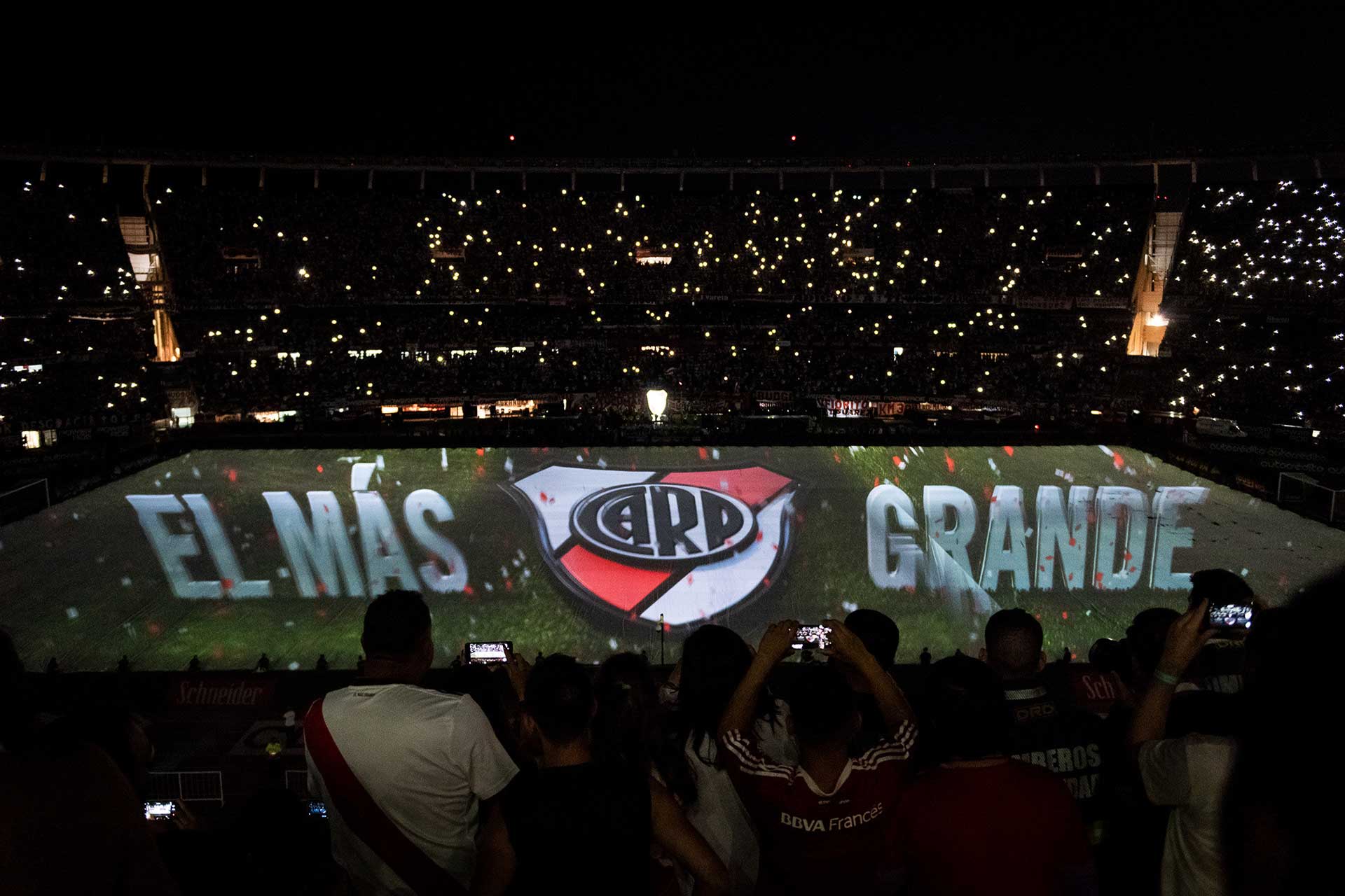 Fotografía de imagen proyectada sobre la manta del campo de juego, en la imagen aparece el escudo de River Plate acompañado de la leyenda 'El Más Grande'. De fondo las luces del estadio apagas y todas las tribunas iluminando con sus celulares, efecto de estrellas. Cobertura del evento '09-12 eterno' organizado por el Club Atlético River Plate