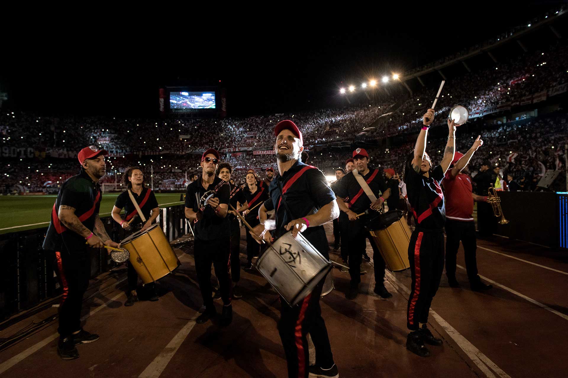 Fotografía del grupo de percusiones 'Choque Urbano' recorriendo la pista de atletismo mientras tocan sus instrumentos y observan las tribunas colmadas. Todos visten una camiseta negra con una banda roja en diagonal desde el hombro hasta la cintura. Cobertura del evento '09-12 eterno' organizado por el Club Atlético River Plate