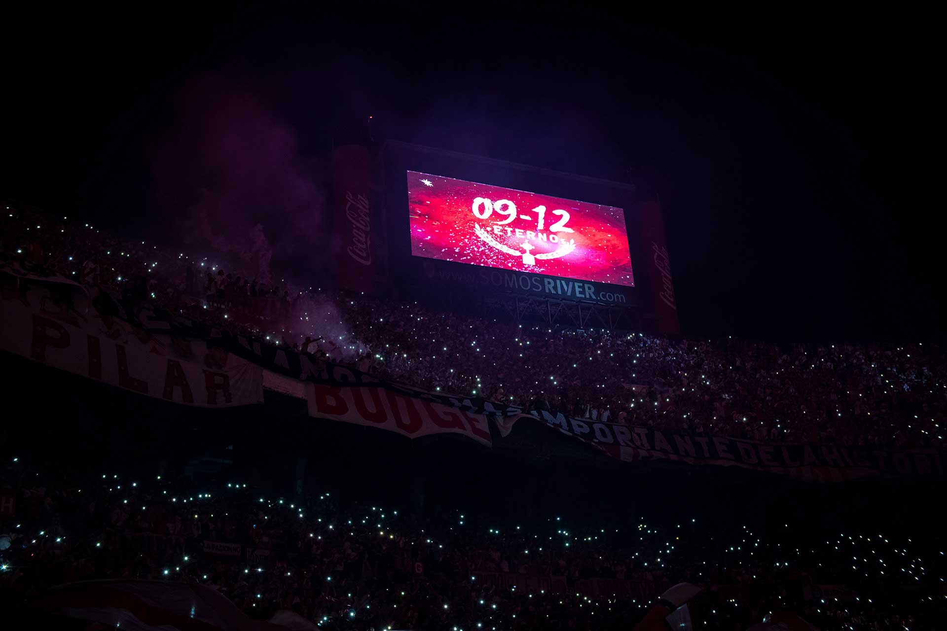 Fotografía de la pantalla del estadio que proyecta el afiche del evento, debajo de la misma, las tribunas del estadio con los hinchas iluminando con sus celulares. Cobertura del evento '09-12 eterno' organizado por el Club Atlético River Plate