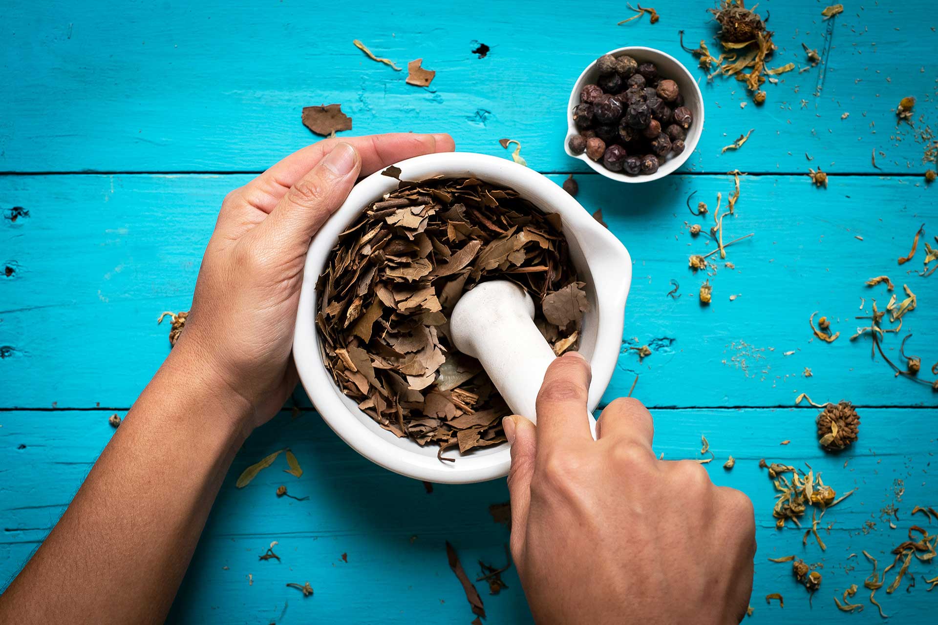 Fotografía de las manos de una persona utilizando un mortero. De fondo la tabla, color turquesa, de una mesa y sobre ellas varias hojas, flores y guindillas empleadas para realizar cosméticos naturales. Material fotográfico para redes sociales de la marca 'Nativa Biocosmética'