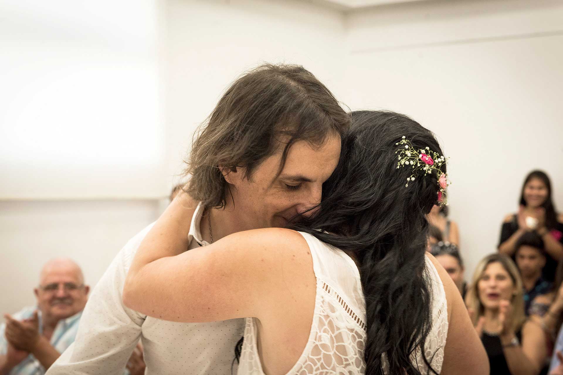 Fotografía de los novios abrazándose emocionados, de fondo los presentes en el Registro Civil. Casamiento de Noe y Ale