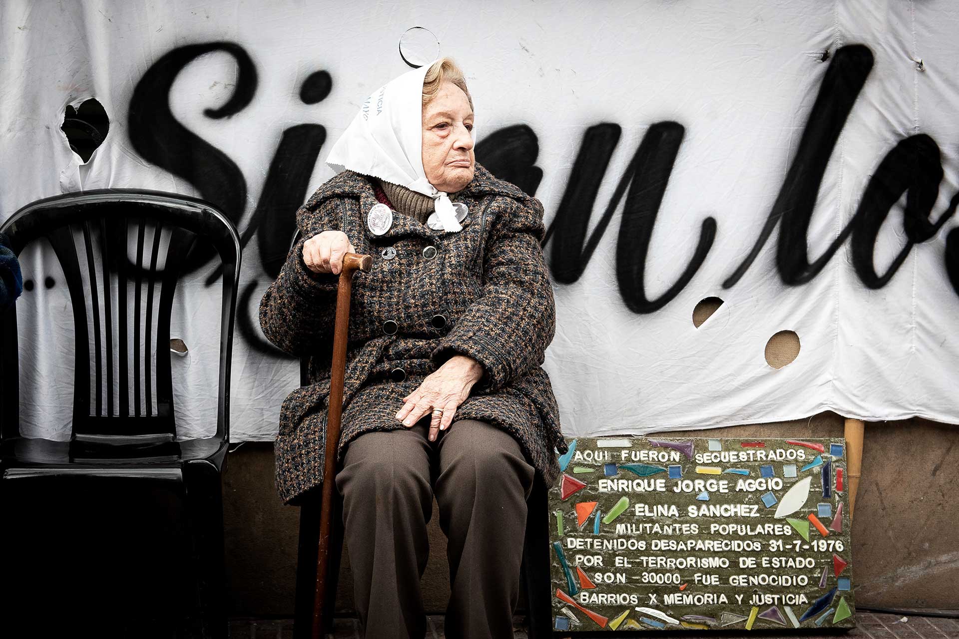 Retrato fotográfico de una 'Abuela de Plaza de Mayo' sentada y a su lado una silla vacía (desaparecido)