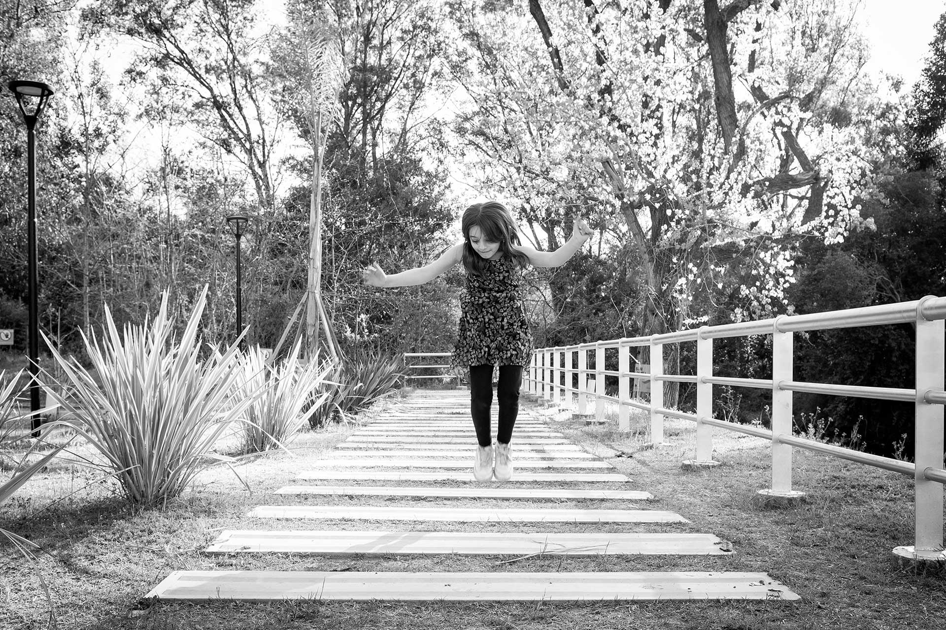 Infancia. Retrato fotográfico, en blanco y negro, de una niña jugando a saltar tablones en un camino de madera