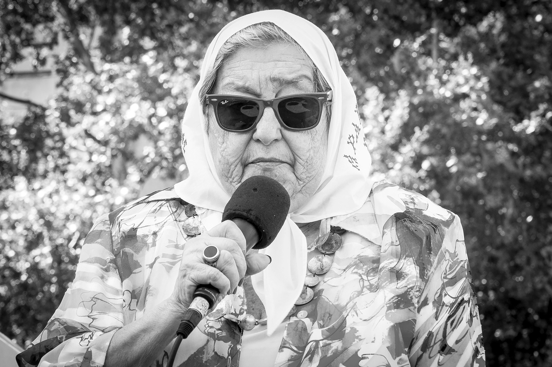 Retrato fotográfico, en blanco y negro, de Hebe de Bonafini, Madre de Plaza de Mayo, sosteniendo un micrófono y reflejándose los icónico pañuelos de 'Madres de Plaza de Mayo'en sus anteojos