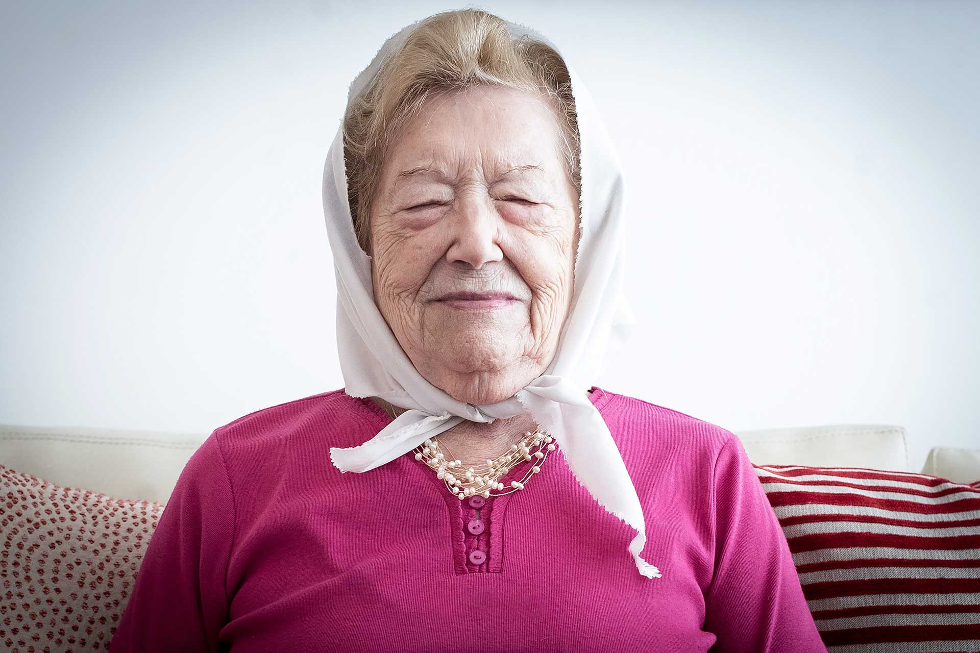 Retrato fotográfico de Sara Rus, Madre de Plaza de Mayo Linea Fundadora, sentada en el sillón de su casa con los ojos cerrados, luego colocarse el icónico pañuelo de 'Madres de Plaza de Mayo'