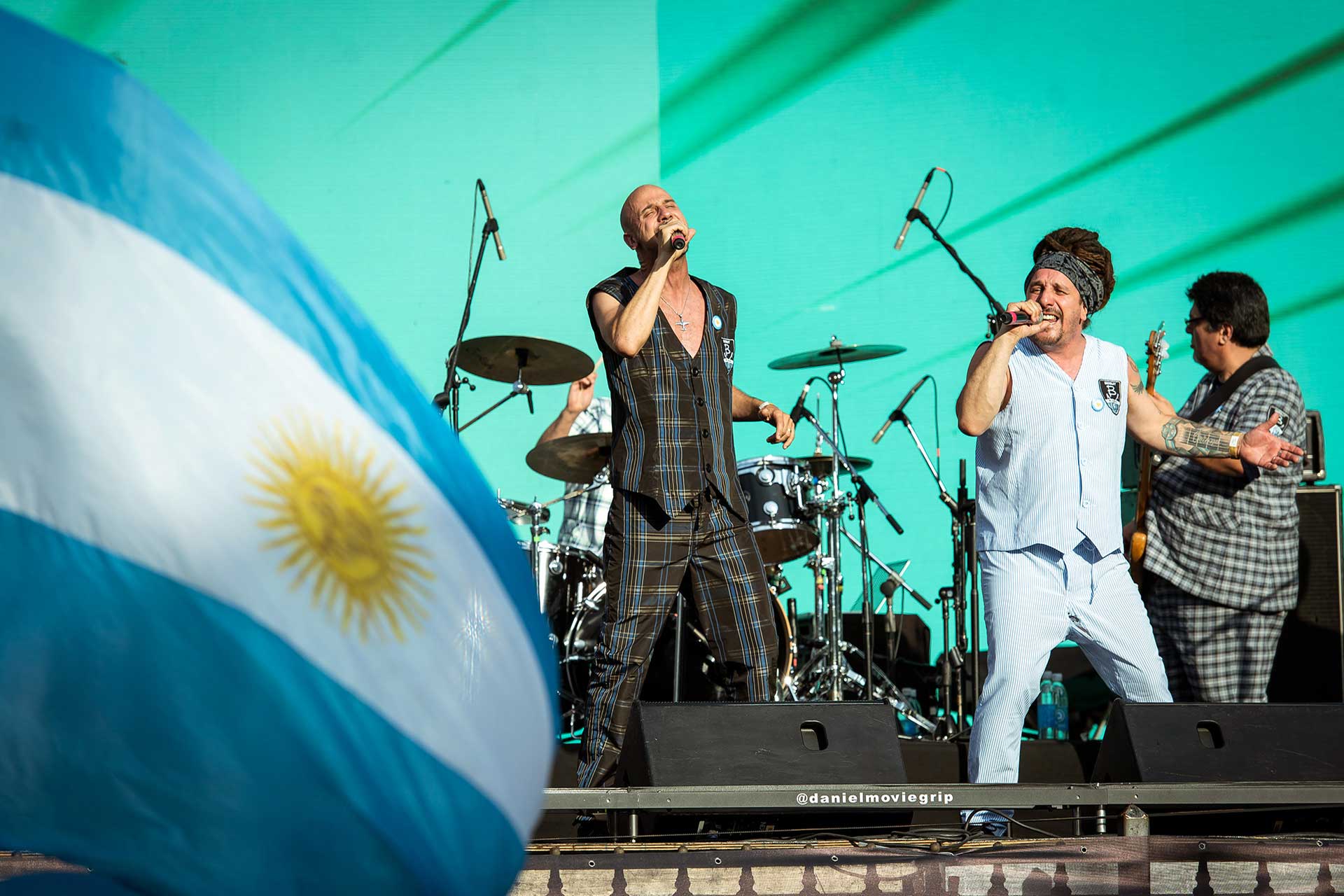 Fotografía de los músicos de Bersuit Vergarabat sobre el escenario, de costado flamea la bandera Argentina