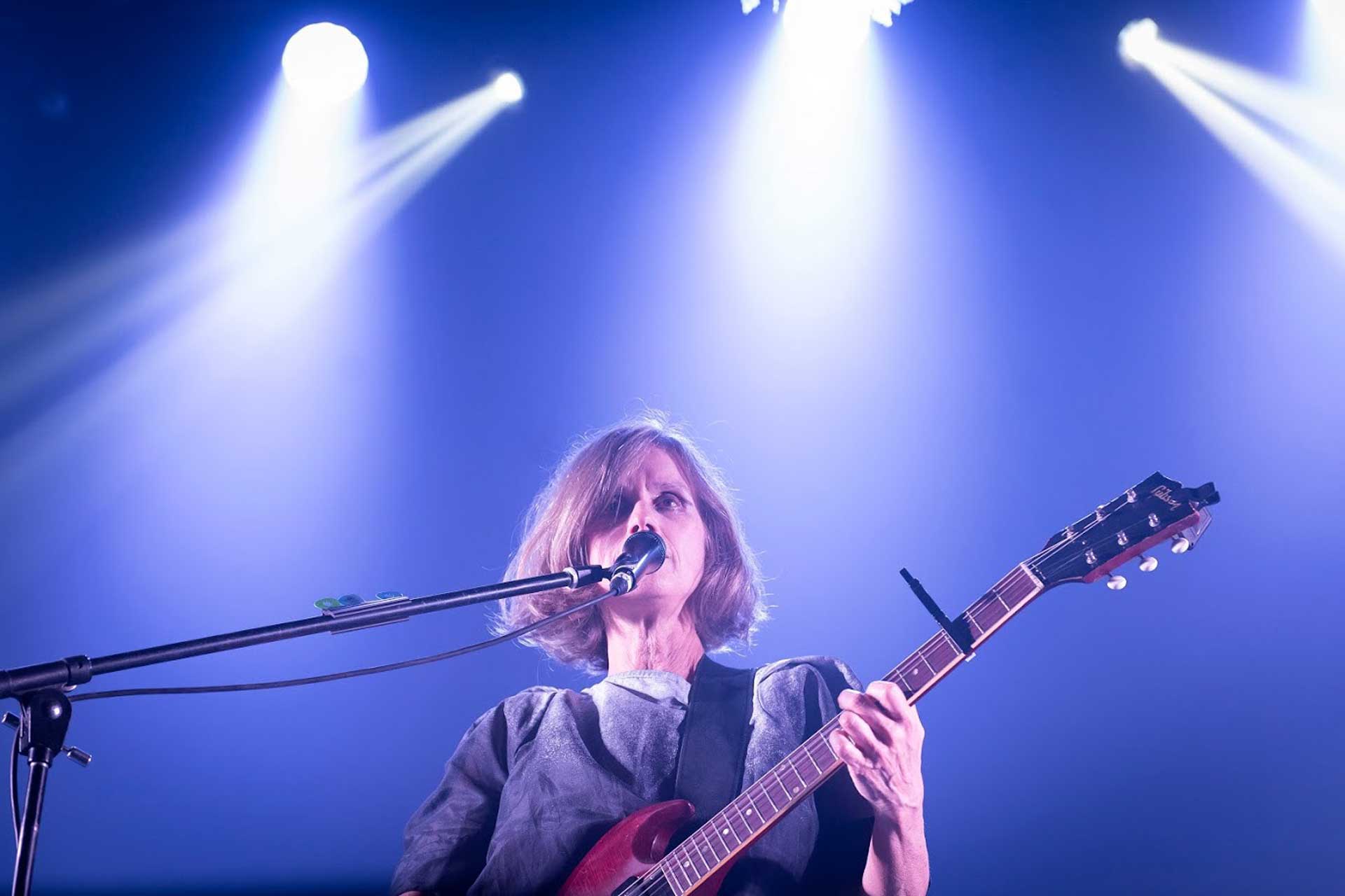 Fotografía de Juana Molina tomada desde el piso del escenario. Aparece ella en primer plano cantando y tocando la guitarra, de fondo juego de luces azules